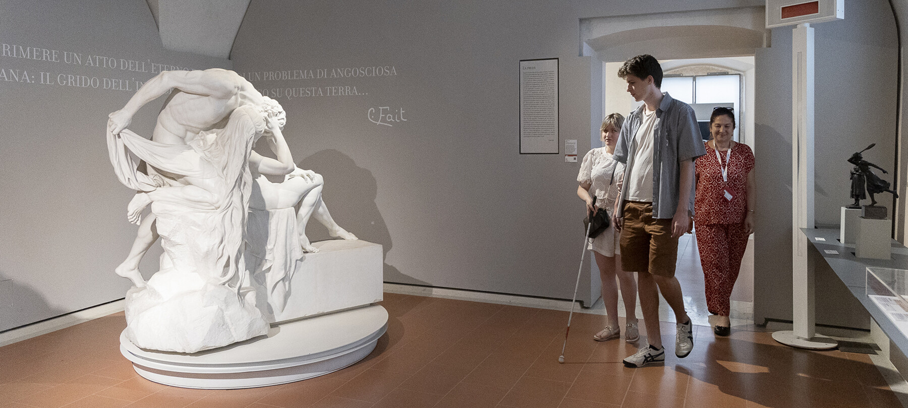 Carlo Fait Hall of the Palazzo Alberti Poja, in Rovereto. On the left is a plaster sculpture depicting a man assaulting and oppressing a woman. Three people enter through the door on the right: a boy, a blind woman, and the tour guide accompanying them. 