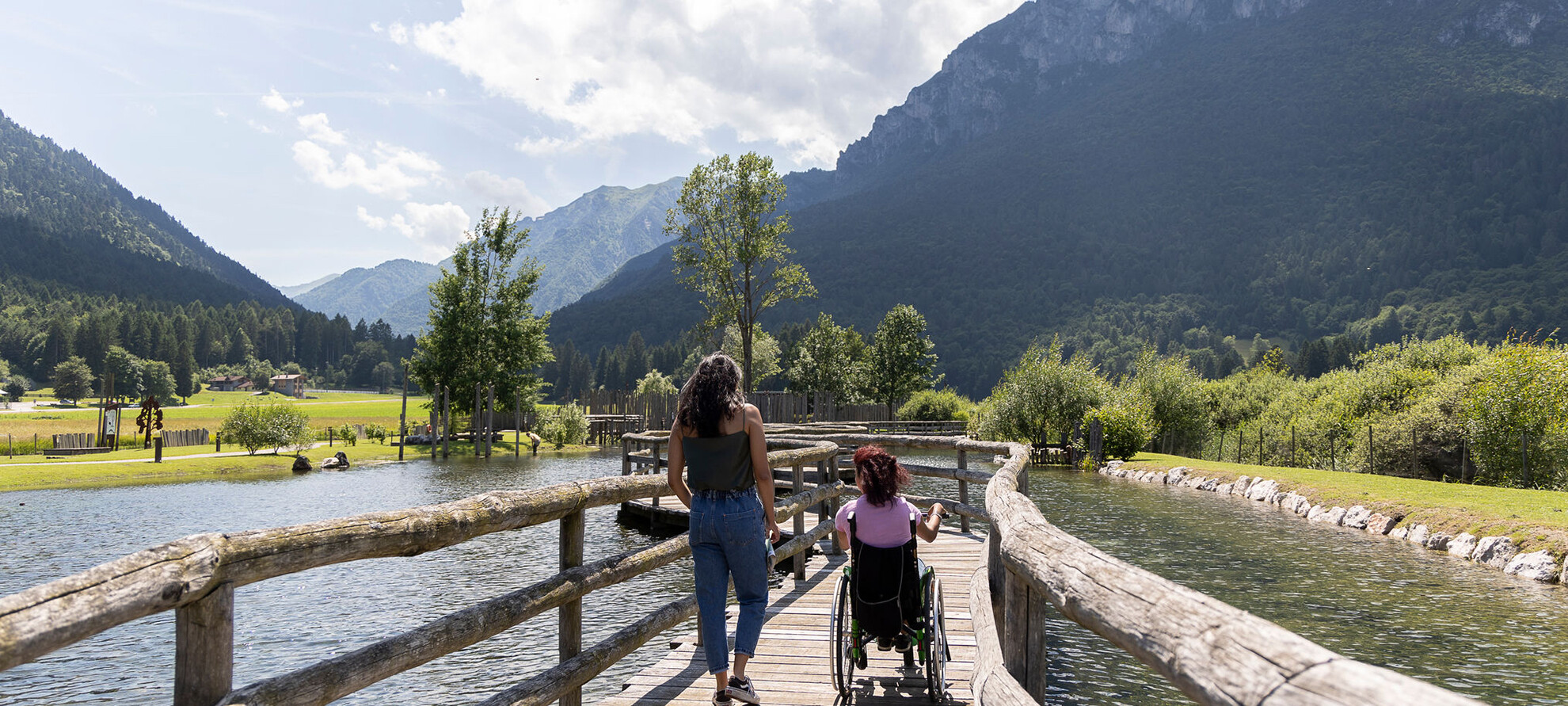 A woman in a wheelchair and a tour guide walk along a catwalk suspended over water. Their backs are turned and they seem to be chatting with each other. It is summer, a beautiful sunny day. The green mountains are reflected on the surface of the water. 