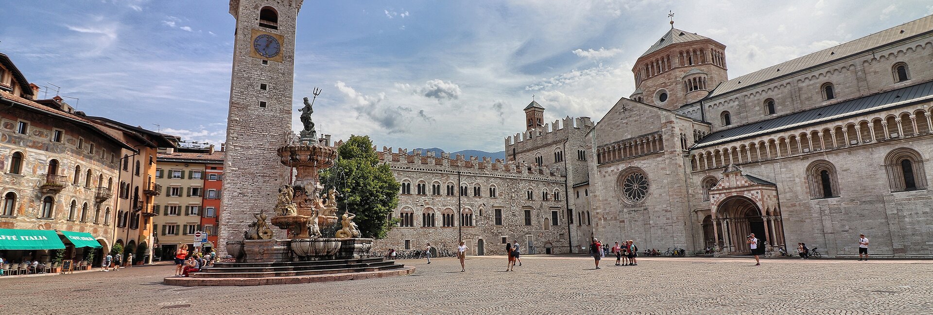 Piazza Duomo a Trento. Sullo sfondo, la Torre Civica, Palazzo Pretorio e la cattedrale di San Vigilio, Duomo della città. Al centro, la fontana del Nettuno. Dietro alla fontana svetta l’unico albero della piazza: un tiglio. La piazza, nell’immagine, è vissuta. Ci sono alcune persone sedute sui gradini della fontana, qualcuno sta scattando delle foto e un gruppetto è in posa davanti alla cattedrale per farsi fotografare. Tutti indossano abiti estivi. Il cielo azzurro è striato dal bianco di nuvole leggere.