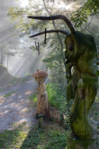 Bosco Arte Stenico, in una giornata di sole in cui i raggi creano giochi di luce attraversando i rami degli alberi. Sul lato del sentiero, alla destra dell’immagine, due statue in legno. Quella in primo piano, alta forse quanto un uomo, potrebbe essere una chimera: volto di civetta, lunghe corna che somigliano alle antenne di una formica, il corpo che pare avvolto in una tunica. La statua in secondo piano, invece, è più piccola e ricorda un torsolo di mela con un corto picciolo in cima.