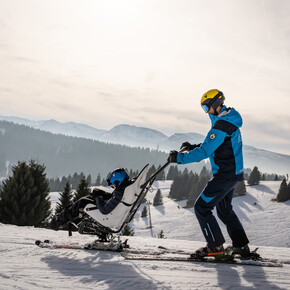 Sci inclusivo in Alpe Cimbra. Una persona con disabilità motoria scia grazie al bi-sci e al supporto di un istruttore specializzato. La luce del sole è morbida e le ombre sulla neve iniziano ad allungarsi: il tramonto non dev’essere lontano.  Nel video, persone con diverse disabilità vivono l’Alpe Cimbra sperimentando varie attività outdoor. Appassionati di biking percorrono ciclabili sbarrierate, sentieri erbosi e attraversano torrenti grazie a handbike adatte alle avventure fuori strada. Chi ama gli sport invernali, invece, si dedica alle attività sulla neve grazie ad attrezzature adatte alle necessità delle persone disabili e agli istruttori specializzati che li accompagnano nelle discese. 