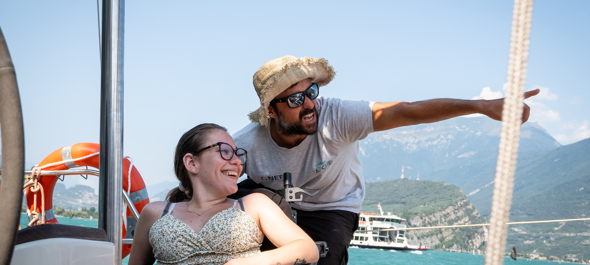 Una donna in carrozzina attraversa il lago di Garda su un’imbarcazione adatta a ospitare persone con disabilità motoria. Accanto a lei, un uomo con gli occhiali da sole e un cappello di paglia indica qualcosa in lontananza. Ha un’espressione buffa che ricorda quella di un pirata: lei ride mentre segue con lo sguardo il punto lontano che lui le sta chiedendo di osservare. | © ©Trentino Marketing_Gianluca Prati_Accessibilita_Riva del Garda_2024_00039
