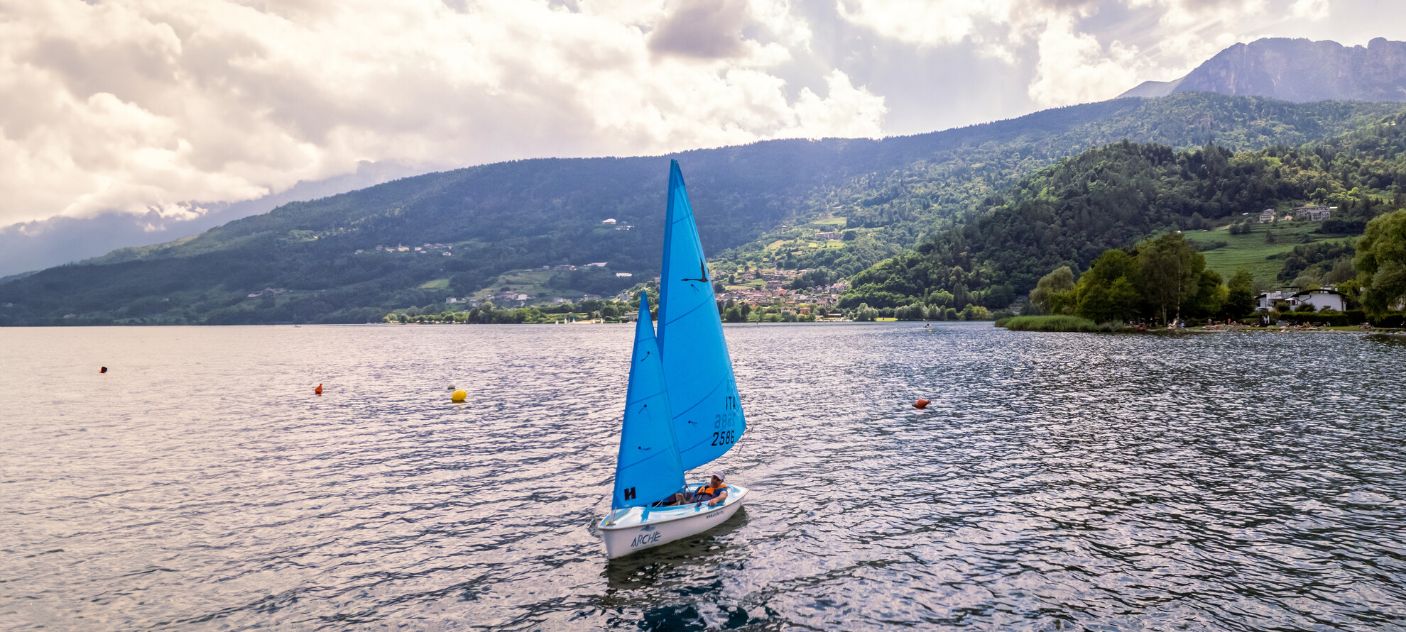 Un’imbarcazione a vela della Cooperativa Arché attraversa il lago di Caldonazzo. È una deriva accessibile adatta anche a persone con disabilità motorie. Non ci sono altre barche nel lago e l’immagine trasmette una sensazione di calma e silenzio. | © ©Trentino Marketing_Gianluca Prati_Accessibilita_Lago di Caldonazzo_2024_00005