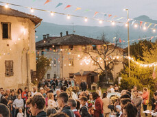 A warm and festive atmosphere fills the streets of Zambana Vecchia during Antiche Terre. The band plays surrounded by people in vintage clothing, under string lights and colorful flags. The community gathers to celebrate tradition and memory.