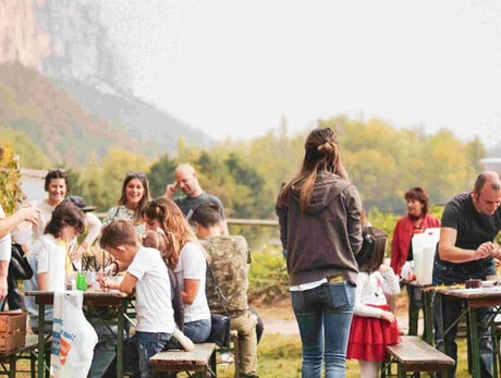 A group of children and adults take part in an outdoor activity surrounded by nature, with craft tables, games, and a warm, communal atmosphere. The event feels friendly and relaxed, ideal for a local gathering. Hills and vineyards can be seen in the background.