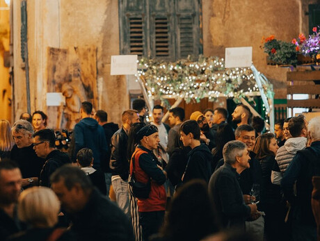 The photo shows a lively outdoor party in a historic village, illuminated by hanging lights and floral decorations. People are chatting and enjoying the convivial atmosphere, with stalls and decorated arches. The ancient walls and wooden shutters add rustic charm.