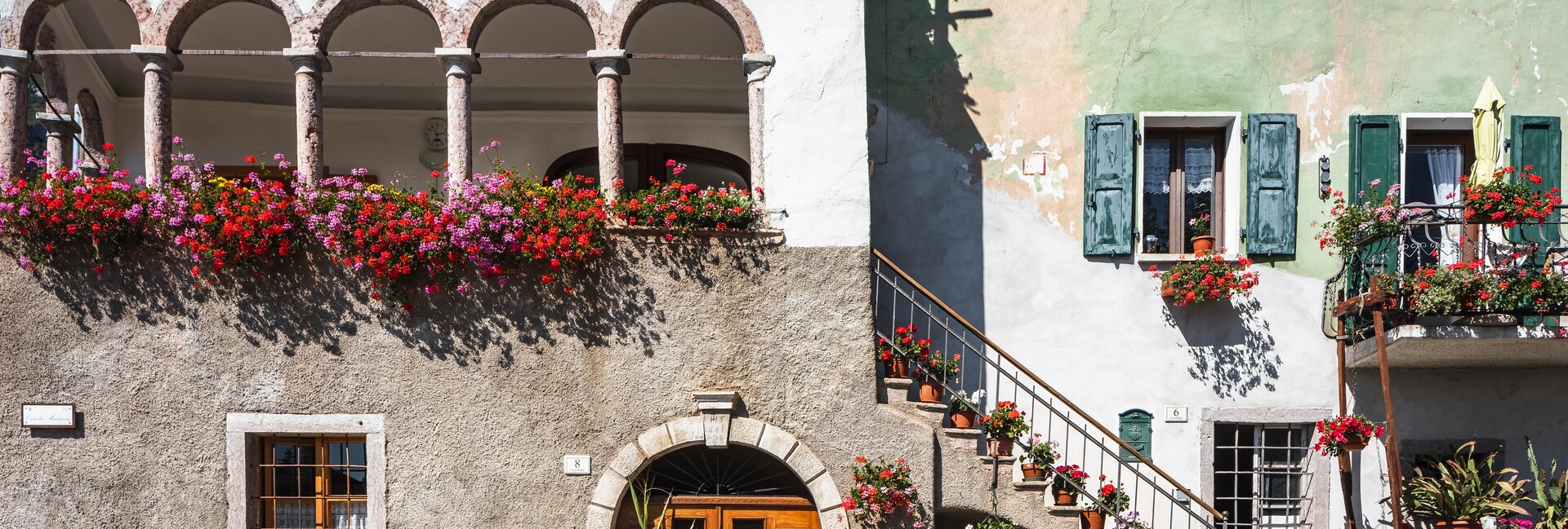 A view of San Lorenzo in Banale on a sunny day. The red and purple spots of the many pots of flowers, perhaps geraniums, stand out in the picture: they are hanging from the windows and the edge of a terrace surrounded by arches, leaning on the steps and on a small balcony. The sunshine and these bright colours convey a sense of cheerfulness.