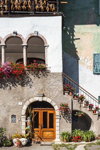 A view of San Lorenzo in Banale on a sunny day. The red and purple spots of the many pots of flowers, perhaps geraniums, stand out in the picture: they are hanging from the windows and the edge of a terrace surrounded by arches, leaning on the steps and on a small balcony. The sunshine and these bright colours convey a sense of cheerfulness.